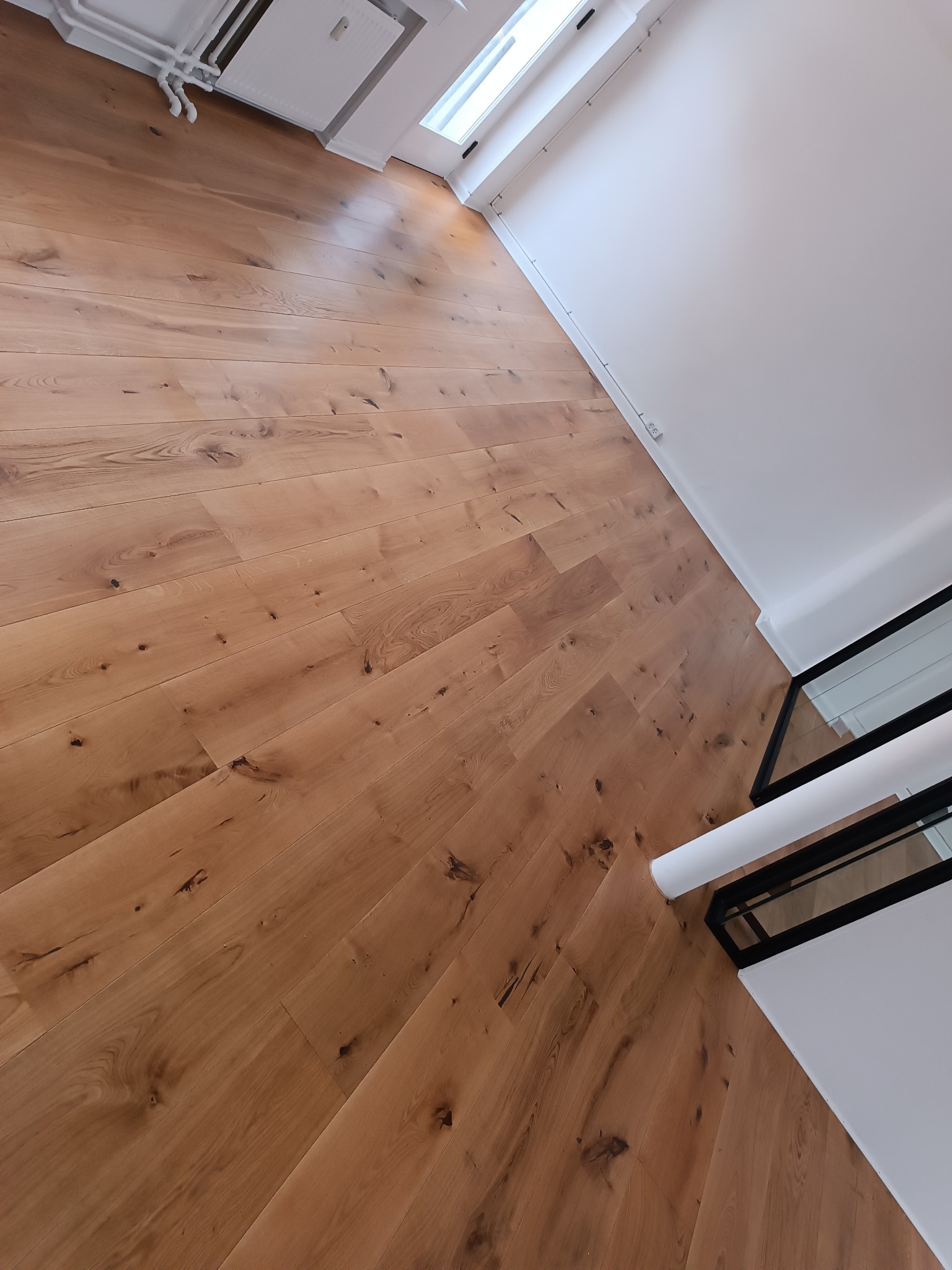 Ułożony parkiet z naturalnego drewna dębowego, widoczne sęki i zróżnicowanie odcieni, jasne ściany i fragment grzejnika w tle.