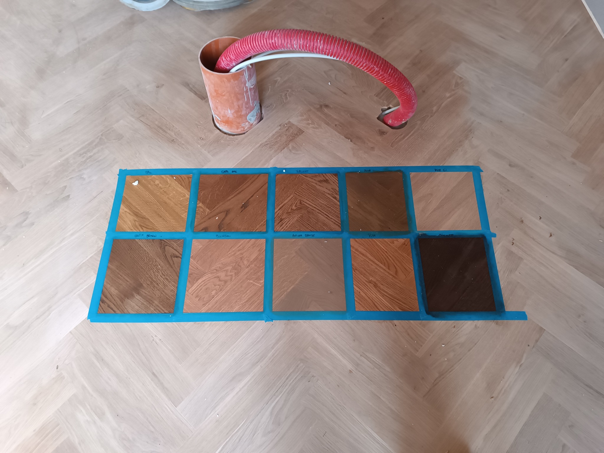 Próbki kolorów parkietu ułożone na podłodze w jodełkę, przyklejone taśmą malarską, widoczna rura wentylacyjna i otwory w podłodze.