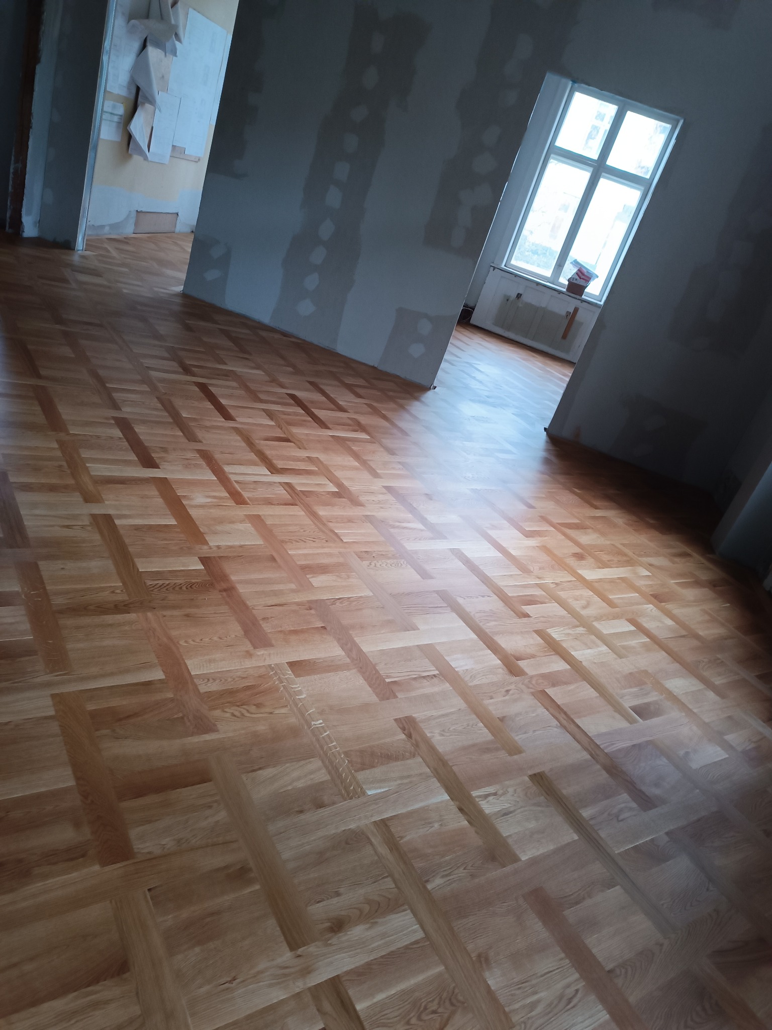 Nowo ułożony parkiet w jodełkę francuską z naturalnego drewna w jasnym pomieszczeniu z widocznymi śladami prac remontowych na ścianach.