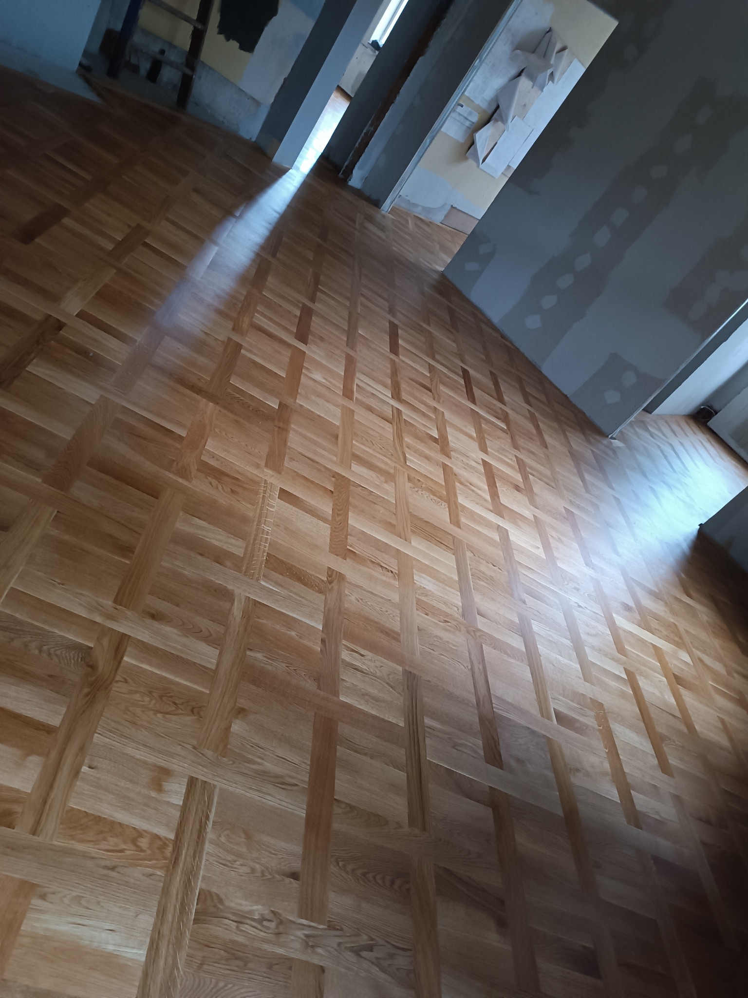 Ukośne ujęcie nowo ułożonego parkietu w jodełkę francuską, widoczne niedokończone ściany i sufit w stanie deweloperskim.