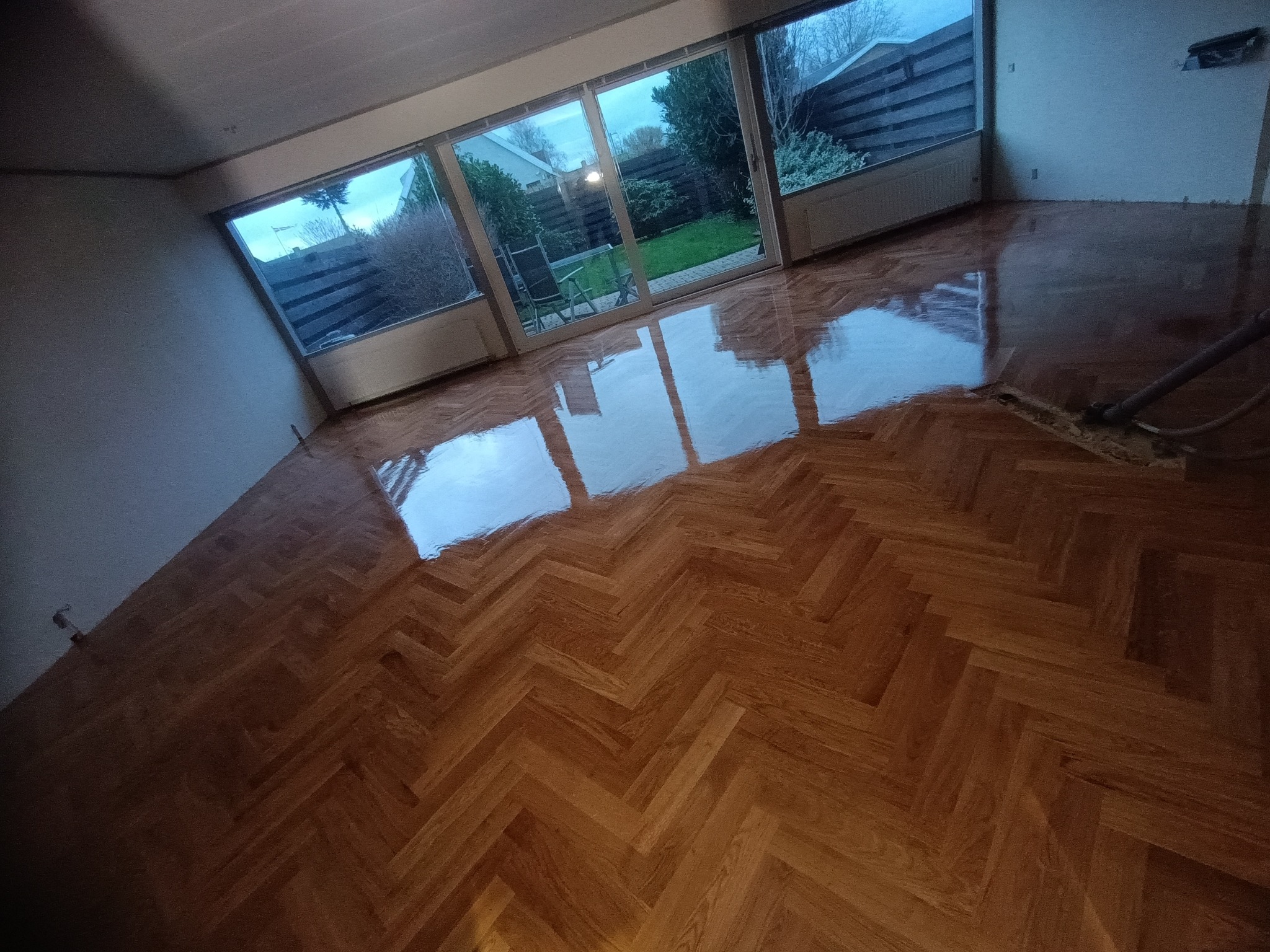 Świeżo położony parkiet w jodełkę, odbijający światło z okien tarasowych z widokiem na ogród, z widocznym fragmentem maszyny do polerowania.