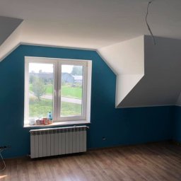 EcoClin - Pomieszczenie po malowaniu ścian na kolor morski, z białym sufitem i oknem. Widoczne drewniane panele podłogowe i grzejnik pod oknem. Gniazdka elektryczne bez osprzętu.