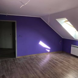 EcoClin - Pokój na poddaszu po malowaniu ścian na fioletowo, z drewnianą podłogą i białym grzejnikiem pod oknem dachowym. Widoczne niedokończone instalacje elektryczne.