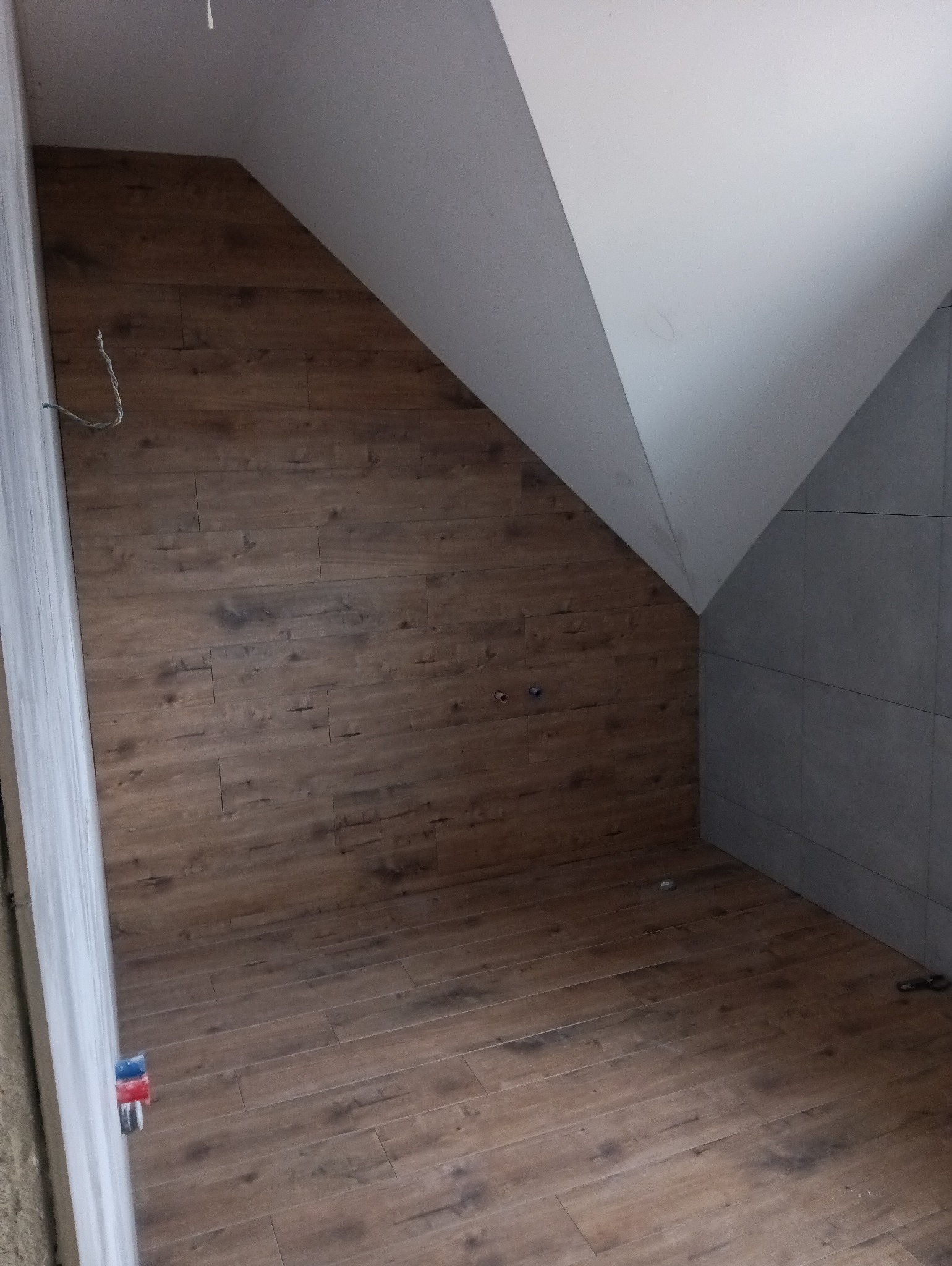 Poddasze po remoncie z drewnopodobnymi panelami na ścianie i podłodze, szare płytki w rogu, widoczne przewody elektryczne i fragment instalacji wodnej.