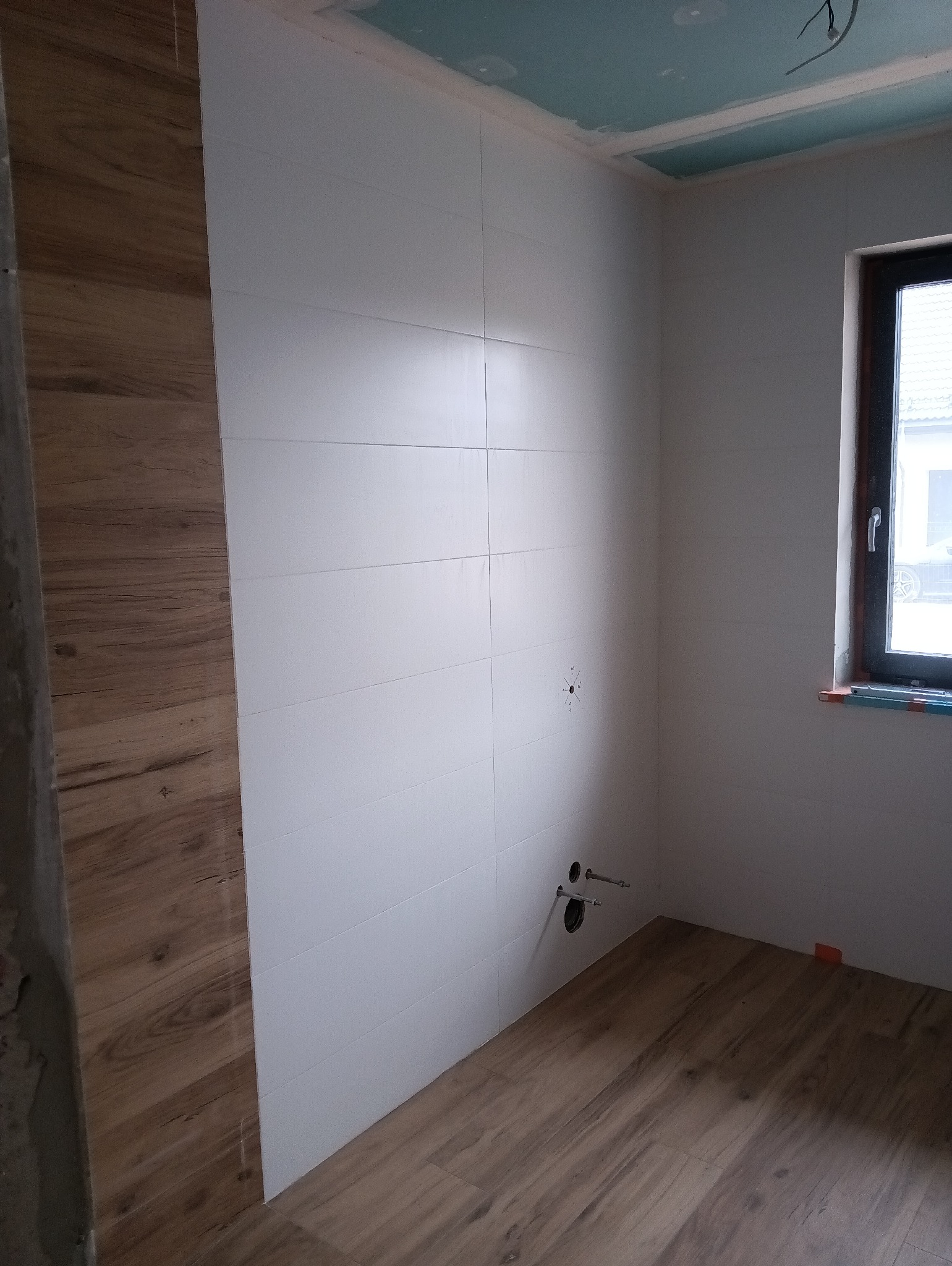Wnętrze łazienki w trakcie wykańczania: częściowo ułożone płytki ścienne w kolorze białym, imitujące poziome panele, oraz podłogowe płytki drewnopodobne, widoczne przyłącza hydrauliczne, okno...