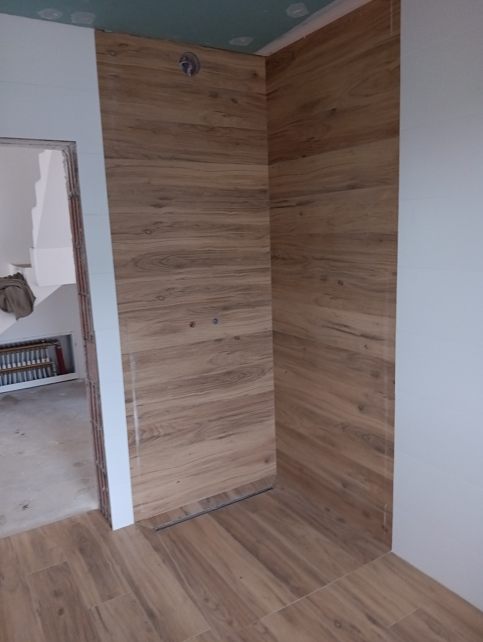 Niedokończona kabina prysznicowa wyłożona panelami imitującymi drewno, widoczne przyłącza wodne, odpływ liniowy i surowy stan wykończenia ścian.