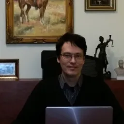 Portret mężczyzny w okularach siedzącego za biurkiem z laptopem, w tle obrazy w złotych ramach i statuetka Temidy