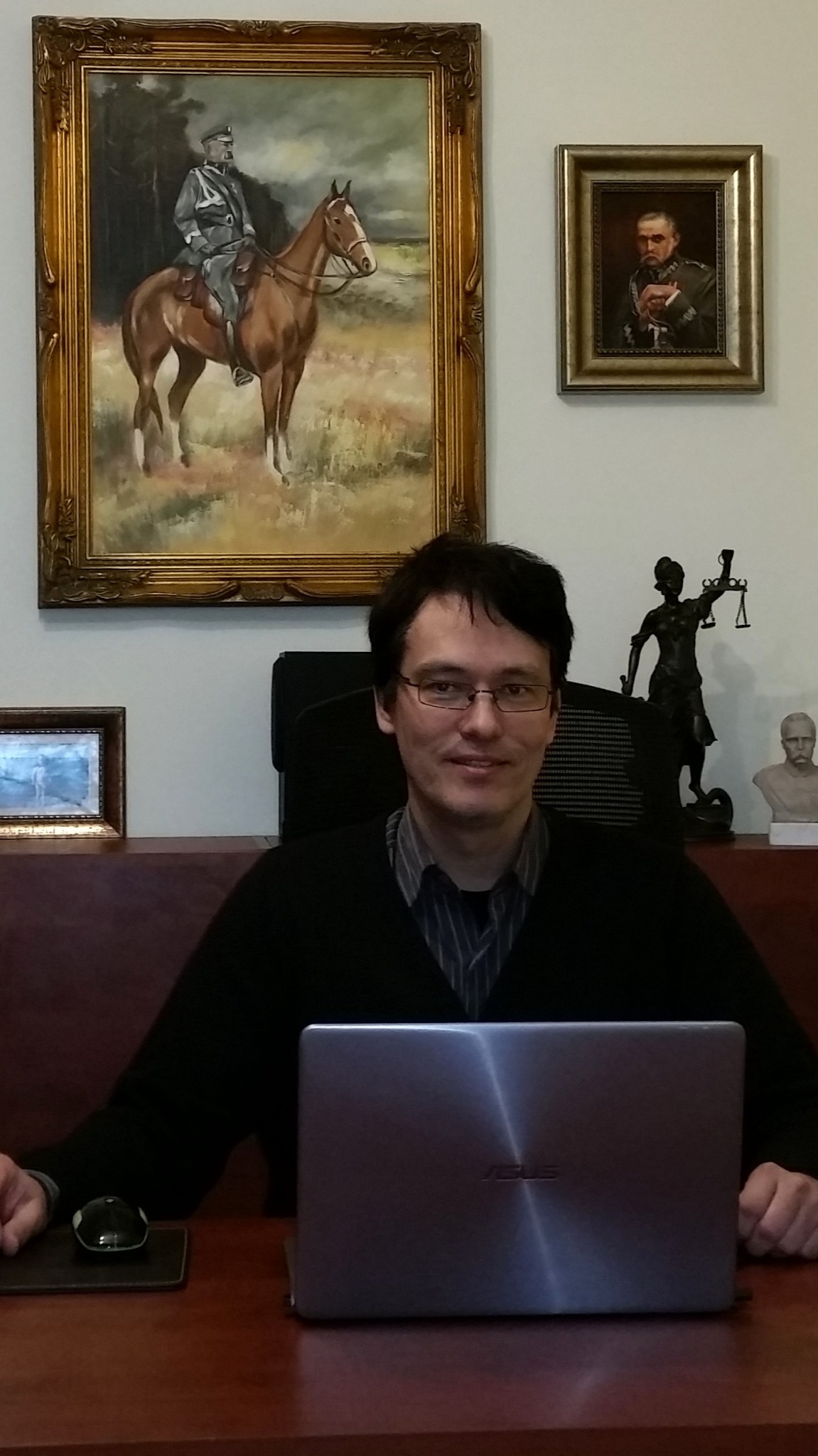 Portret mężczyzny w okularach siedzącego za biurkiem z laptopem, w tle obrazy w złotych ramach i statuetka Temidy