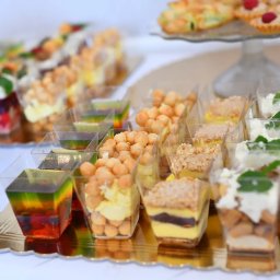 FotoArtNyk - Różnorodne desery w małych, przezroczystych pojemnikach i na szklanych paterach, ułożone na złotej tacy z falistym brzegiem, prezentowane na białym obrusie.