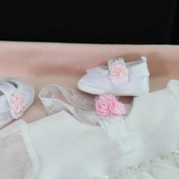 FotoArtNyk - Białe buciki niemowlęce z różowymi różyczkami, leżące obok białej sukienki z koronkowym wykończeniem i opaską z różowymi różami na różowym tle.