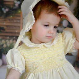 FotoArtNyk - Portret małej dziewczynki w żółtej sukience i czepku z koronką, zamyślona mina, ręka na głowie, tło z motywami wiosennymi.