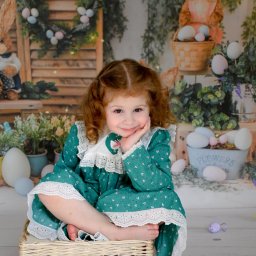 FotoArtNyk - Uśmiechnięta dziewczynka z rudymi kręconymi włosami w zielonej sukience z koronką, siedząca na wiklinowym koszyku na tle dekoracji wielkanocnych z zajączkami i jajkami.