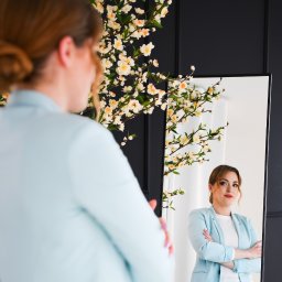 FotoArtNyk - Kobieta w błękitnej marynarce, widziana w lustrze, z dekoracją kwiatową w tle, w eleganckim wnętrzu.