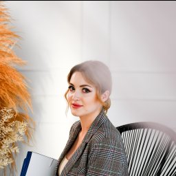 FotoArtNyk - Portret kobiety w kraciastej marynarce siedzącej na plecionym krześle, z teczką w ręku, na jasnym tle z dekoracyjną trawą pampasową.