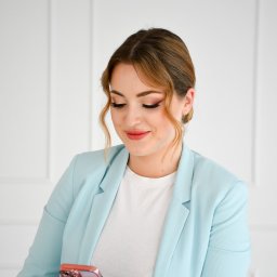 FotoArtNyk - Kobieta w błękitnej marynarce patrzy na smartfon w różowym etui, stojąc na tle białej ściany z subtelnym wzorem paneli.