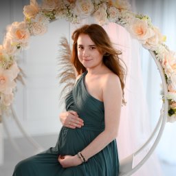 FotoArtNyk - Kobieta w zielonej sukni ciążowej siedzi na huśtawce ozdobionej kwiatami w jasnym studiu.