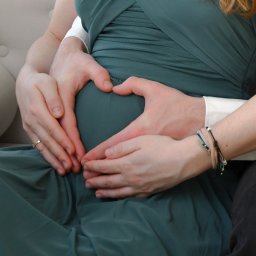 FotoArtNyk - Dłonie kobiety z pierścionkiem i mężczyzny układają się w kształt serca na brzuchu przyszłej mamy w zielonej sukience.