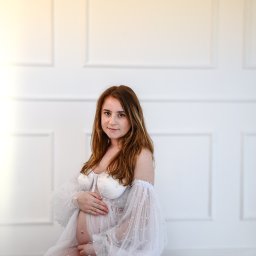 FotoArtNyk - Kobieta w ciąży w zwiewnej, białej sukni z perłami, siedząca na podłodze na tle białej ściany z panelami.