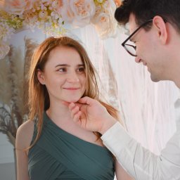 FotoArtNyk - Uśmiechnięta kobieta w zielonej sukience, dotykana przez mężczyznę w okularach na tle dekoracji z jasnych kwiatów i zwiewnych tkanin.