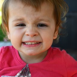 FotoArtNyk - Portret uśmiechniętej, rozzłoszczonej dziewczynki w różowej koszulce z cekinami, pokazującej zęby na rozmytym tle zieleni i szarości.