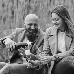 FotoArtNyk - Uśmiechnięta para w plenerze: mężczyzna z brodą nalewa szampana do kieliszków, czarno-biała fotografia.