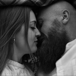 FotoArtNyk - Czarno-białe zbliżenie na profil kobiety z kolczykiem i brodatego mężczyzny, stykających się nosami pod kocem w kratę.