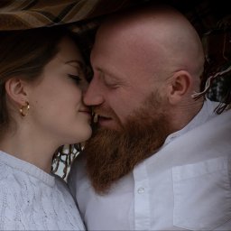 FotoArtNyk - Intymne zbliżenie na parę, kobieta z blond włosami i złotymi kolczykami oraz mężczyzna z rudą brodą, tulą się pod kocem w ciepłym oświetleniu.