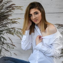 FotoArtNyk - Portret młodej kobiety w okularach, z rozpiętą białą koszulą i dżinsach, siedzącej na tle białych desek i suchych traw.