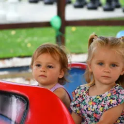 Dwie małe dziewczynki siedzą obok siebie na karuzeli, patrząc w tym samym kierunku, z zielonym trawnikiem i drzewami w tle.