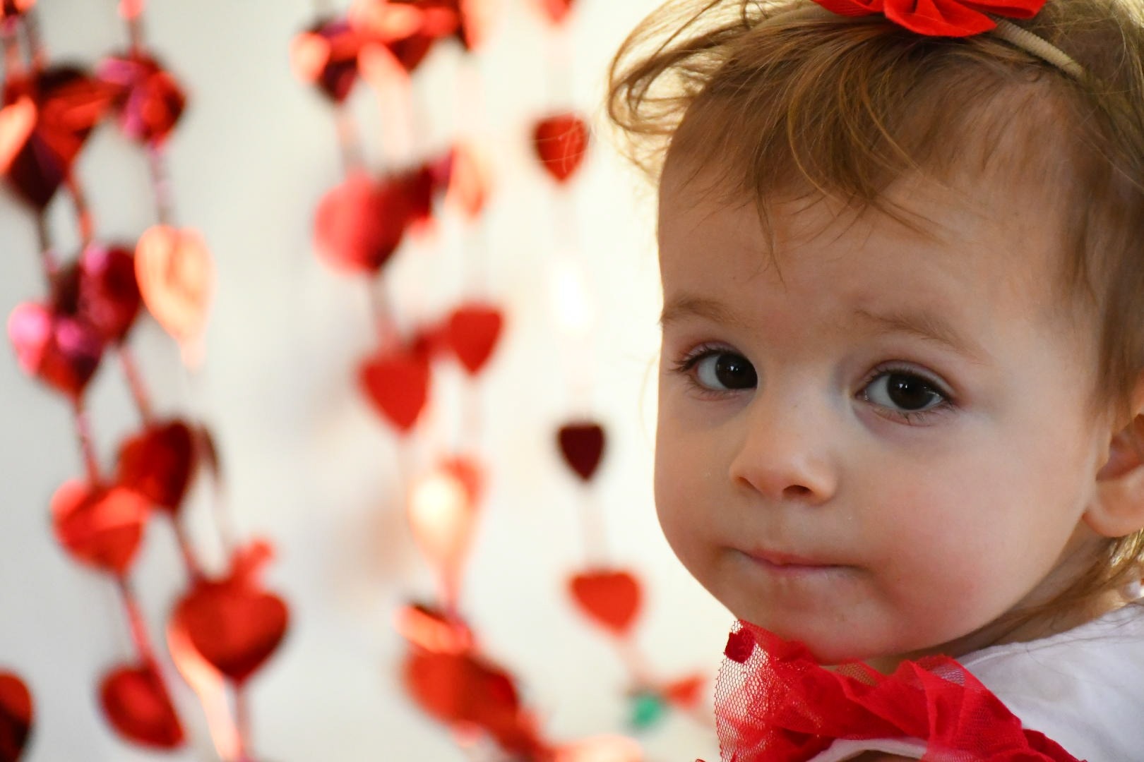 Portret dziewczynki z czerwoną kokardą we włosach i naszyjnikiem z czerwonej siateczki, w tle girlanda z czerwonych serc.