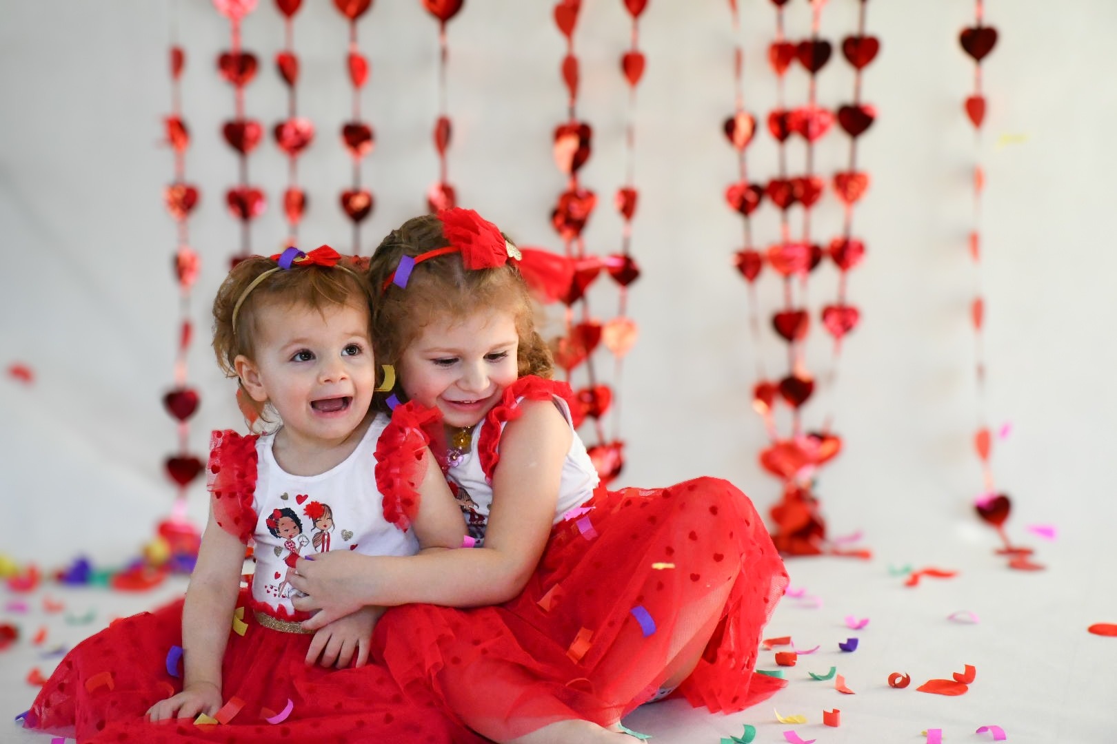 Dwie małe dziewczynki w czerwonych sukienkach i kokardach, siedzą na podłodze z konfetti, na tle girlandy z czerwonych serc.