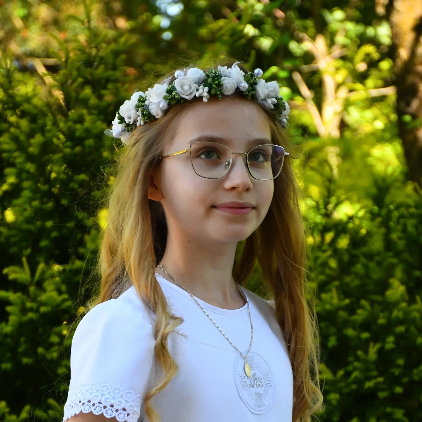 Portret dziewczynki w białej sukience i wianku, z medalikiem IHS na szyi, na tle zieleni. Ujęcie z bliska, skupiające się na twarzy i stroju komunijnym.