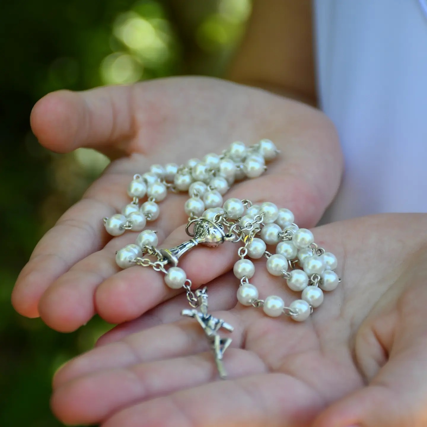 Ręce trzymają biały różaniec z perłami i srebrnym krzyżykiem na tle zieleni.