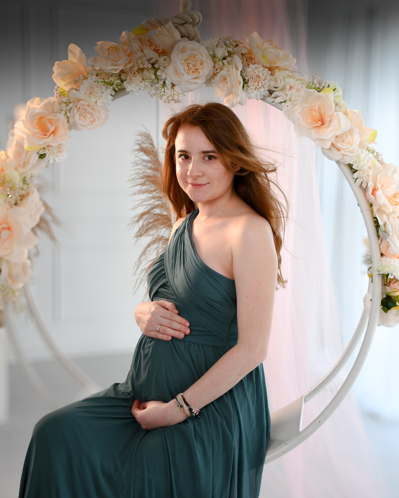 Kobieta w zielonej sukni ciążowej siedzi na huśtawce ozdobionej kwiatami w jasnym studiu.