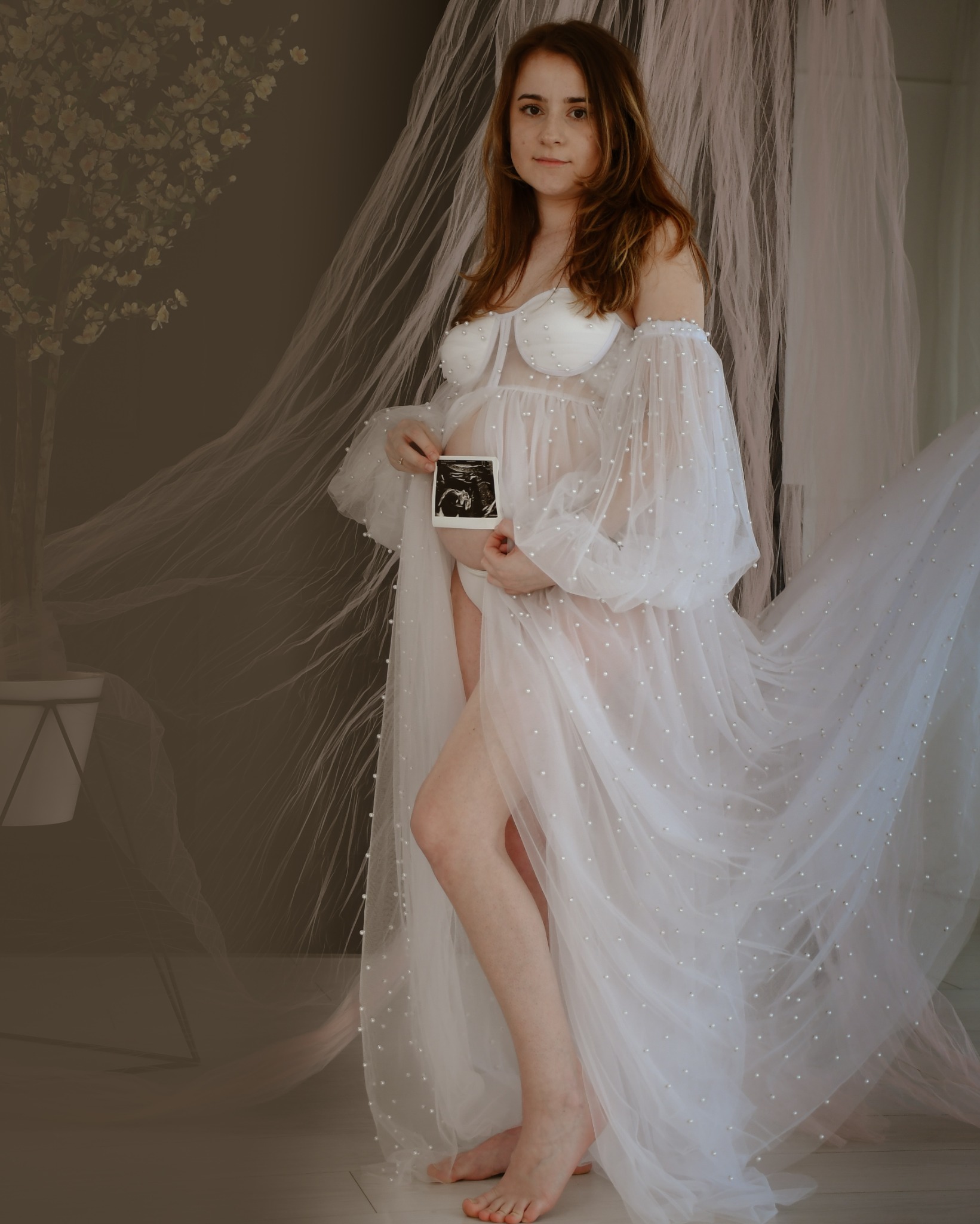 Kobieta w zwiewnej, białej sukni z perłami, trzymająca zdjęcie USG na tle delikatnej zasłony i dekoracji kwiatowej.
