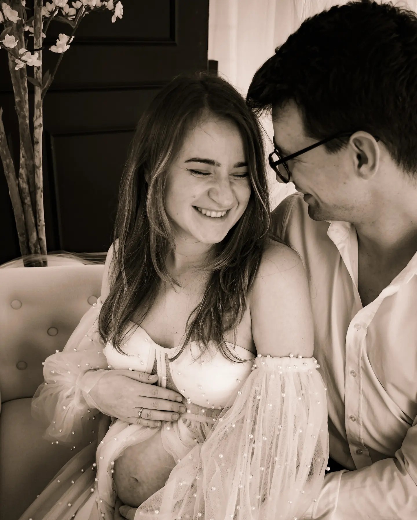 Czarno-białe zdjęcie uśmiechniętej kobiety w ciąży w jasnej sukience z perłami, obejmowanej przez mężczyznę w okularach, siedzących na kremowej sofie z dekoracyjnym drzewkiem w tle.