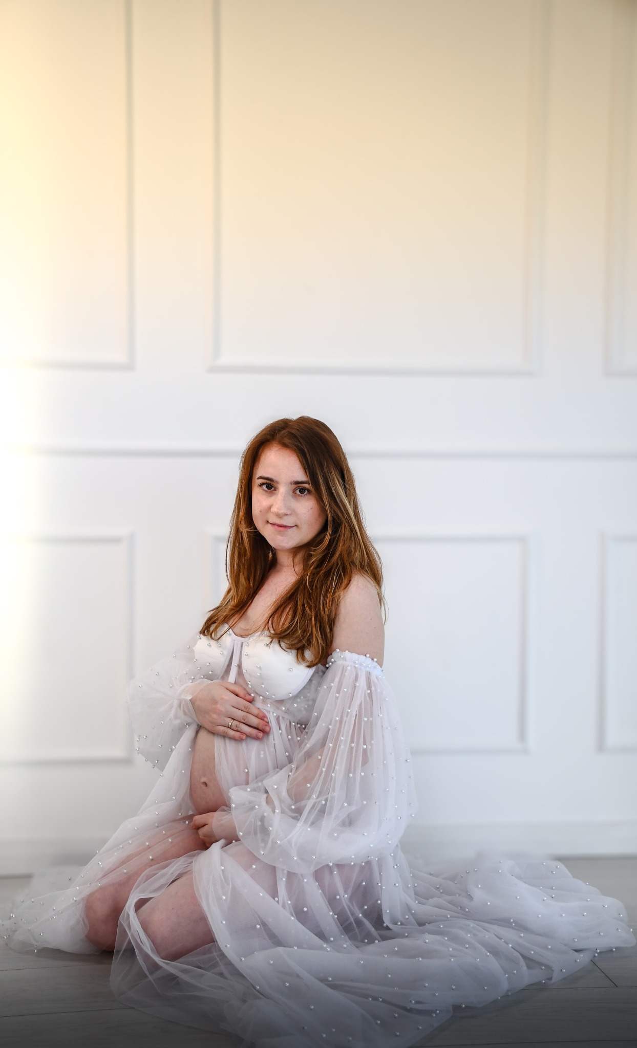 Kobieta w ciąży w zwiewnej, białej sukni z perłami, siedząca na podłodze na tle białej ściany z panelami.