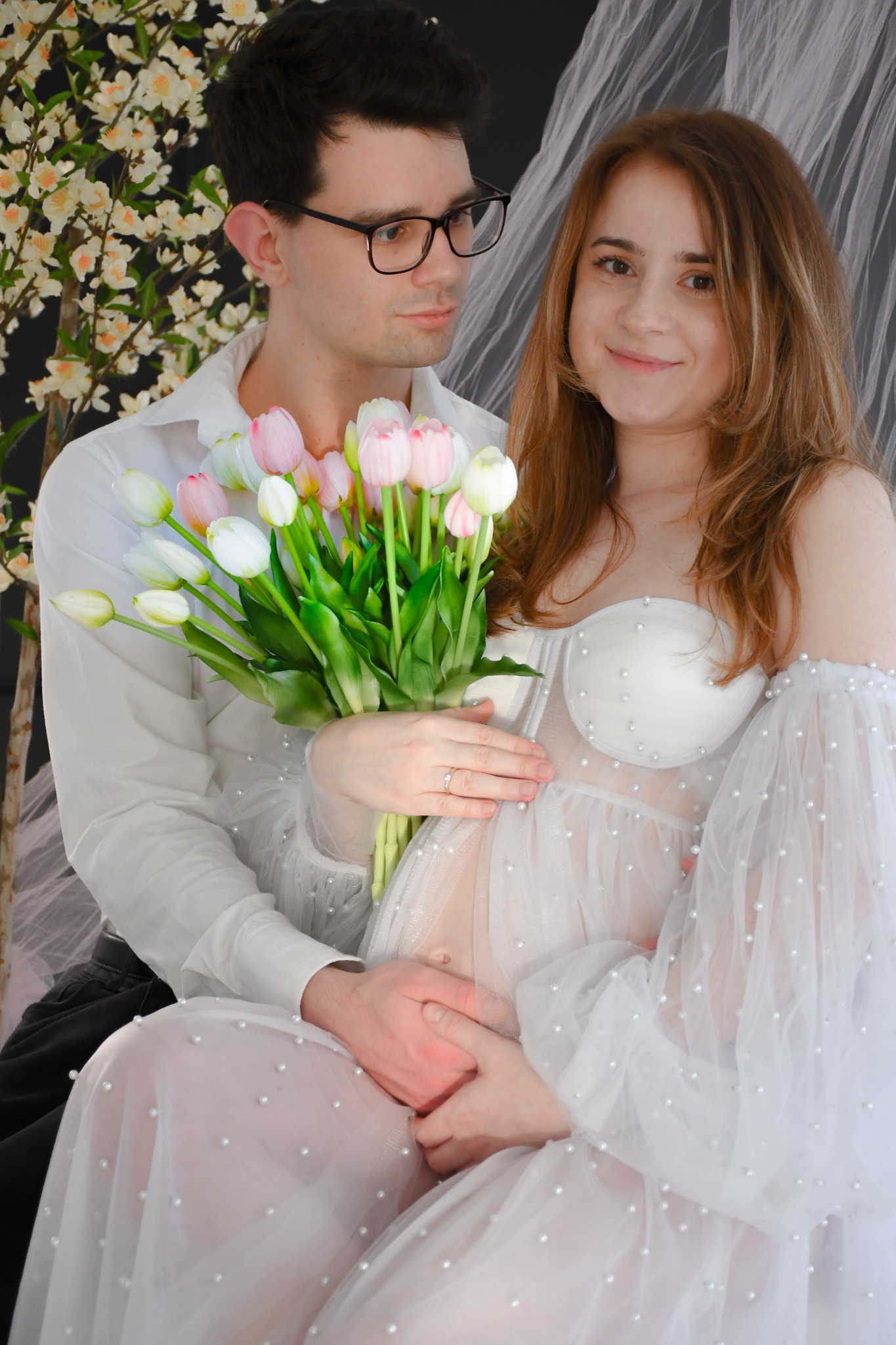 Uśmiechnięta kobieta w zwiewnej, białej sukni z perełkami, trzymająca bukiet tulipanów, pozuje z mężczyzną w okularach na tle kwitnącego drzewa.