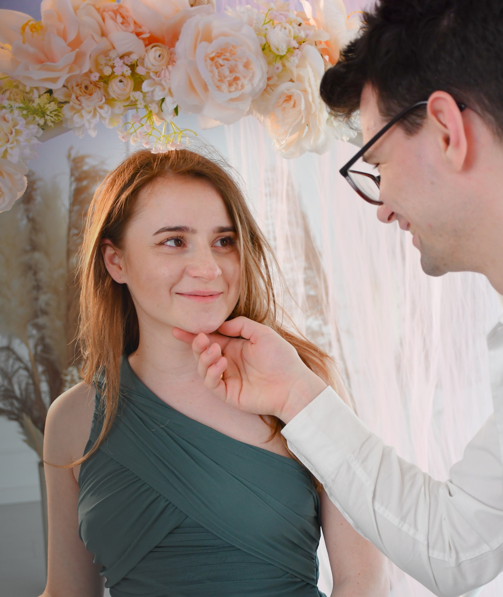 Uśmiechnięta kobieta w zielonej sukience, dotykana przez mężczyznę w okularach na tle dekoracji z jasnych kwiatów i zwiewnych tkanin.
