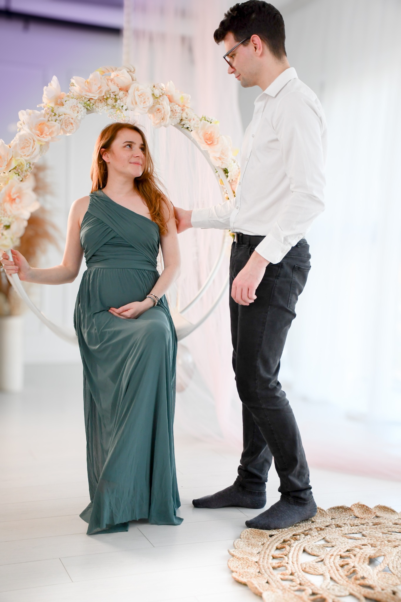 Kobieta w długiej zielonej sukni siedzi na huśtawce z kwiatami, obok stoi mężczyzna w białej koszuli i czarnych spodniach, spoglądają na siebie w jasnym wnętrzu.