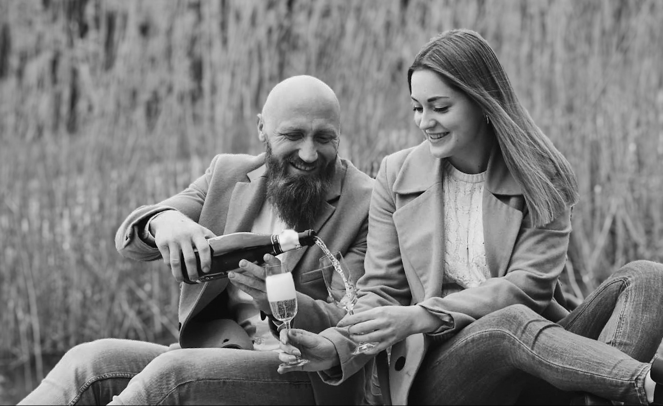 Uśmiechnięta para w plenerze: mężczyzna z brodą nalewa szampana do kieliszków, czarno-biała fotografia.