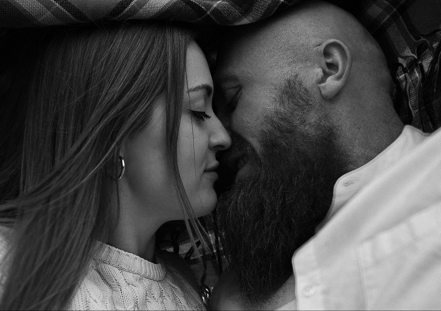 Czarno-białe zbliżenie na profil kobiety z kolczykiem i brodatego mężczyzny, stykających się nosami pod kocem w kratę.