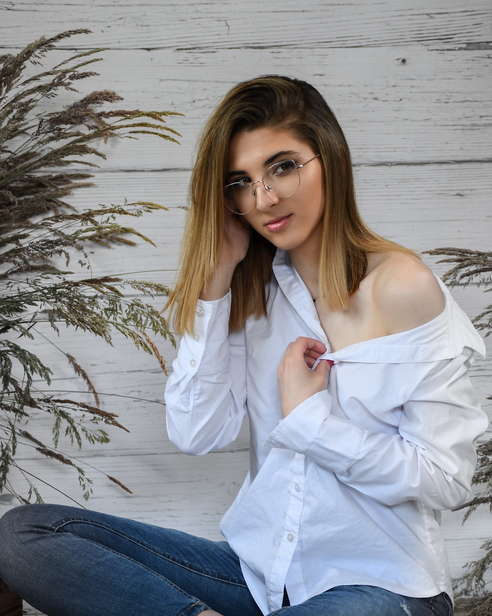 Portret młodej kobiety w okularach, z rozpiętą białą koszulą i dżinsach, siedzącej na tle białych desek i suchych traw.