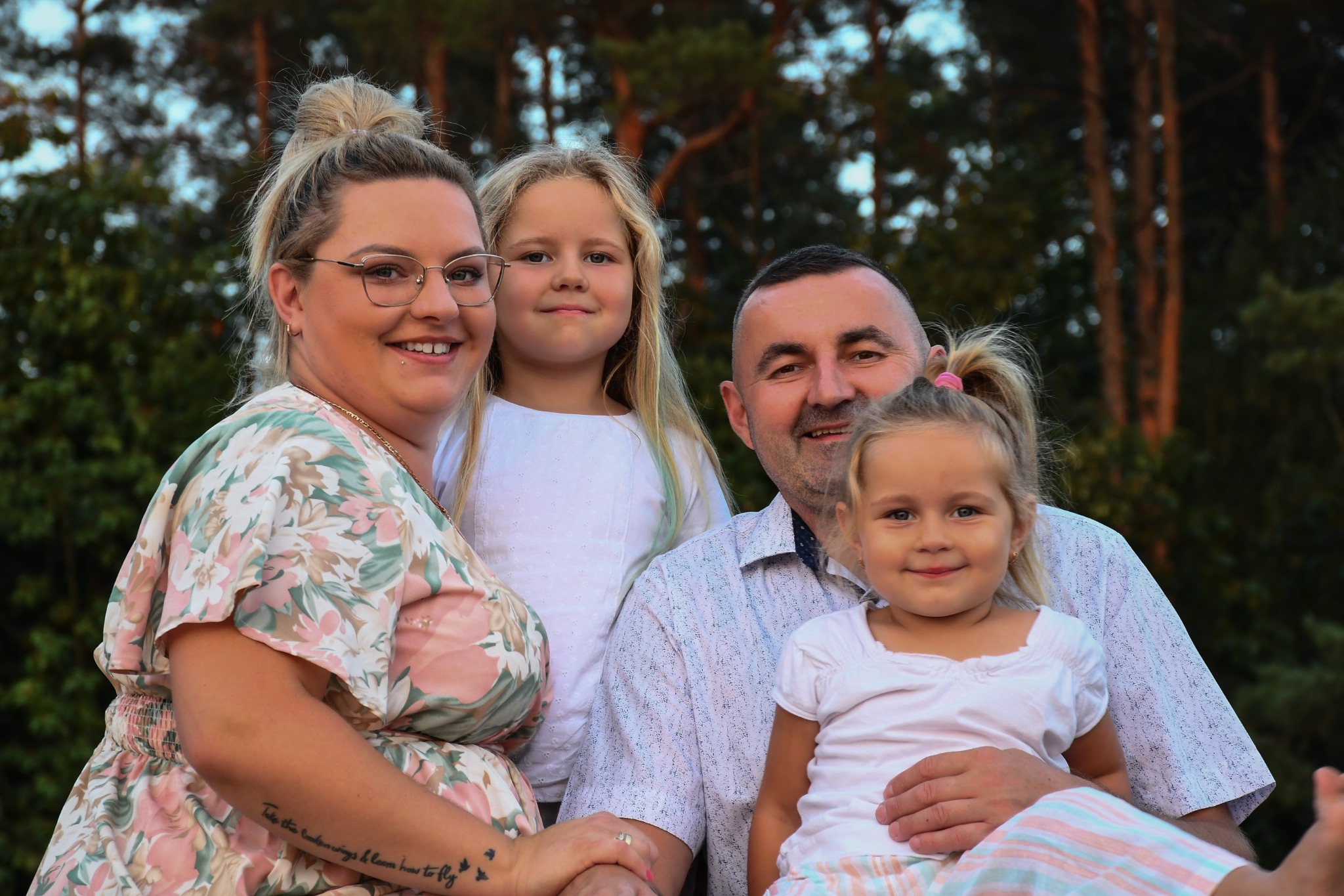 Portret uśmiechniętej rodziny: mama w kwiecistej sukience z tatuażem na ramieniu, tata z dwiema córkami na tle zieleni.