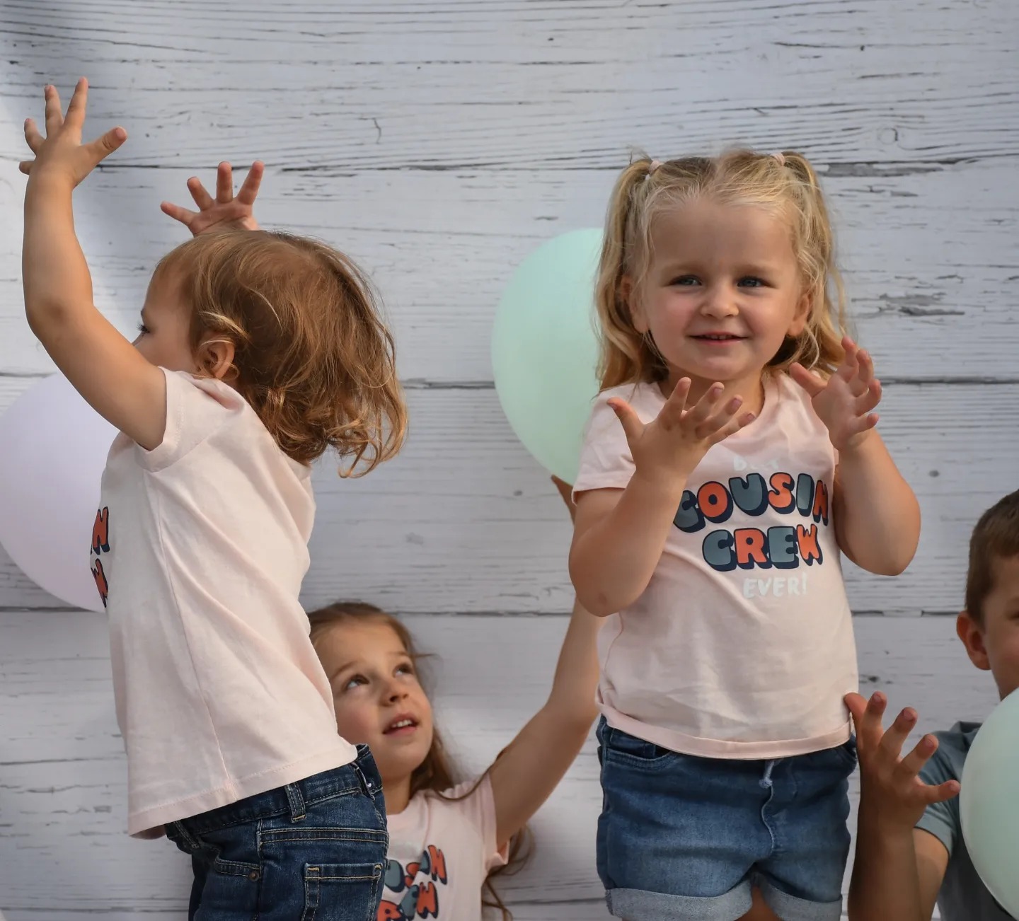 Grupa roześmianych dzieci w jasnych ubraniach, pozujących z balonami na tle białej, drewnianej ściany.
