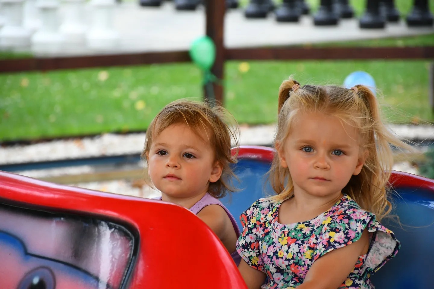 Dwie małe dziewczynki siedzą obok siebie na karuzeli, patrząc w tym samym kierunku, z zielonym trawnikiem i drzewami w tle.