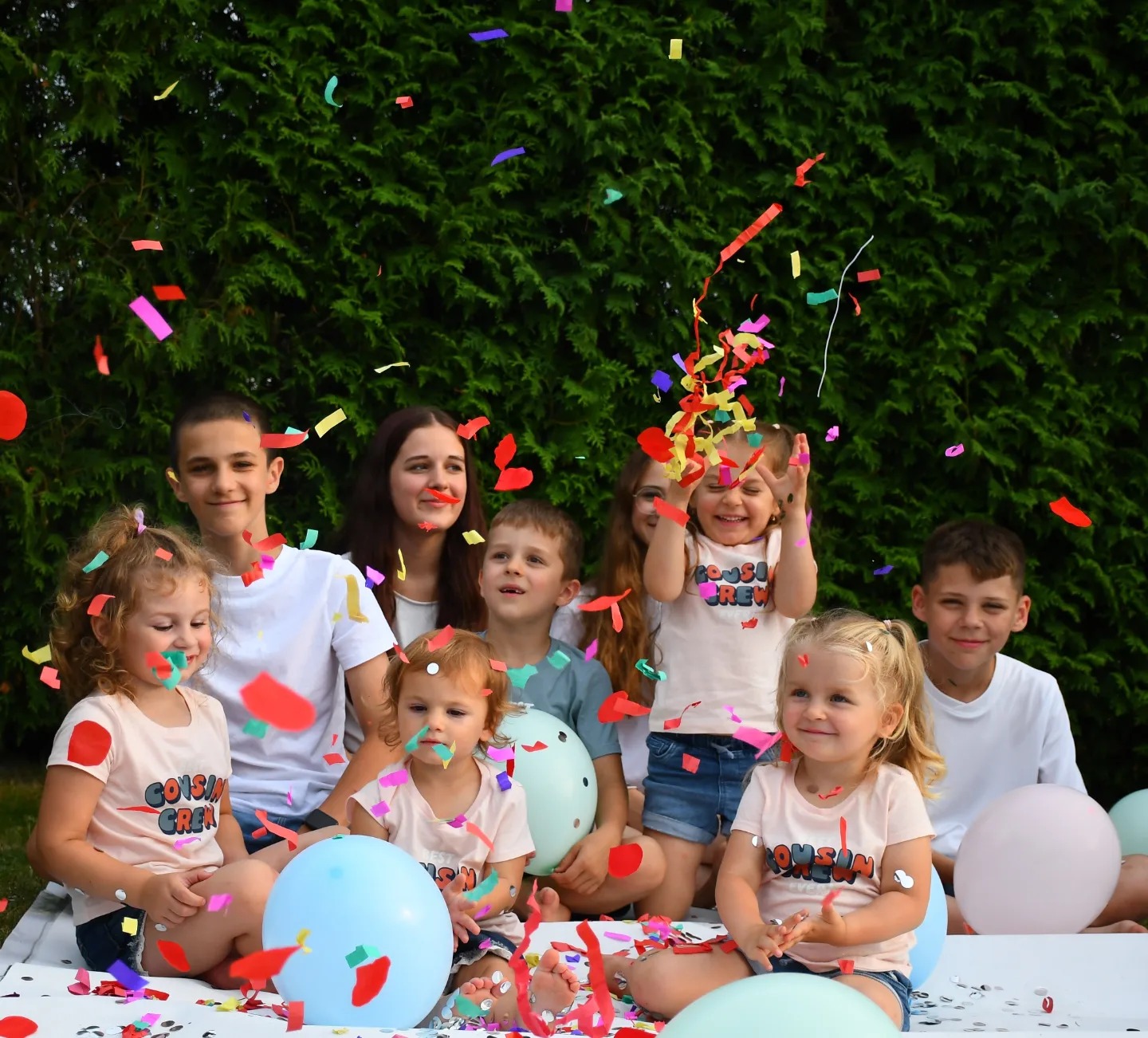 Grupa dzieci w koszulkach z napisem 'Cousin Crew' siedzi na białym tle z balonami, podczas gdy kolorowe konfetti opada z góry na tle zielonego żywopłotu.