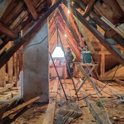 Jakub Tylka - Poddasze w trakcie prac remontowych, widoczne drewniane krokwie, fragment ściany, narzędzia i przewody elektryczne, majster w tle przy oknie.