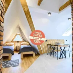 Pokój na poddaszu z dwiema pojedynczymi łóżkami, drewnianą podłogą, małym oknem w kształcie trójkąta, ceglanymi filarami i stołem z krzesłami, całość w jasnej kolorystyce z naklejką 'Good Taste...