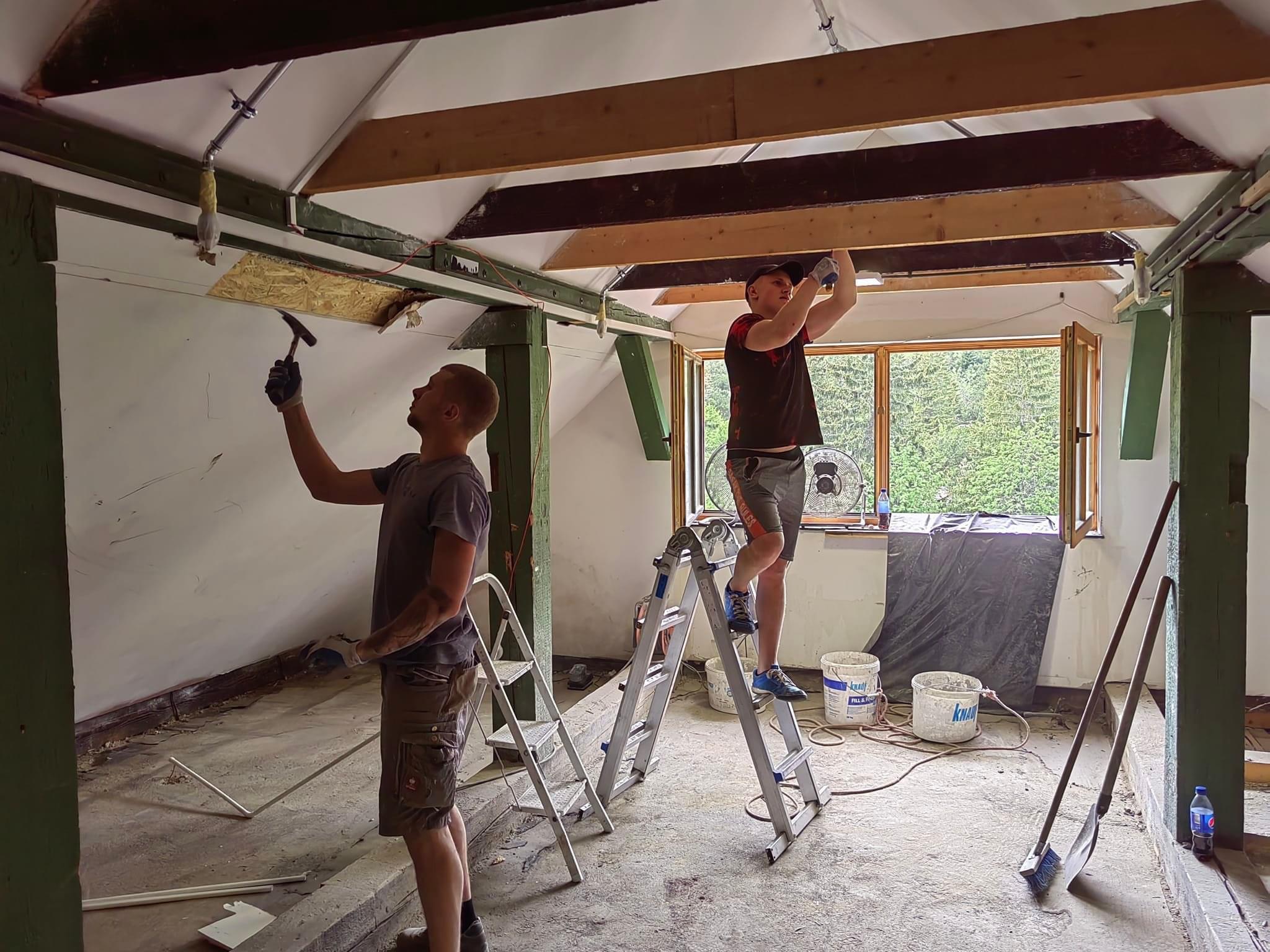 Dwóch mężczyzn na drabinach pracuje nad sufitem poddasza, jeden z młotkiem, drugi mocuje drewnianą belkę, widoczne narzędzia i wiadra z zaprawą.